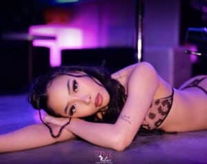 Jeune femme brune en soutien-gorge léopard se repose sur le sol, éclairage rose/violet. Tatouage sur le bras, logo "Pink Palace".