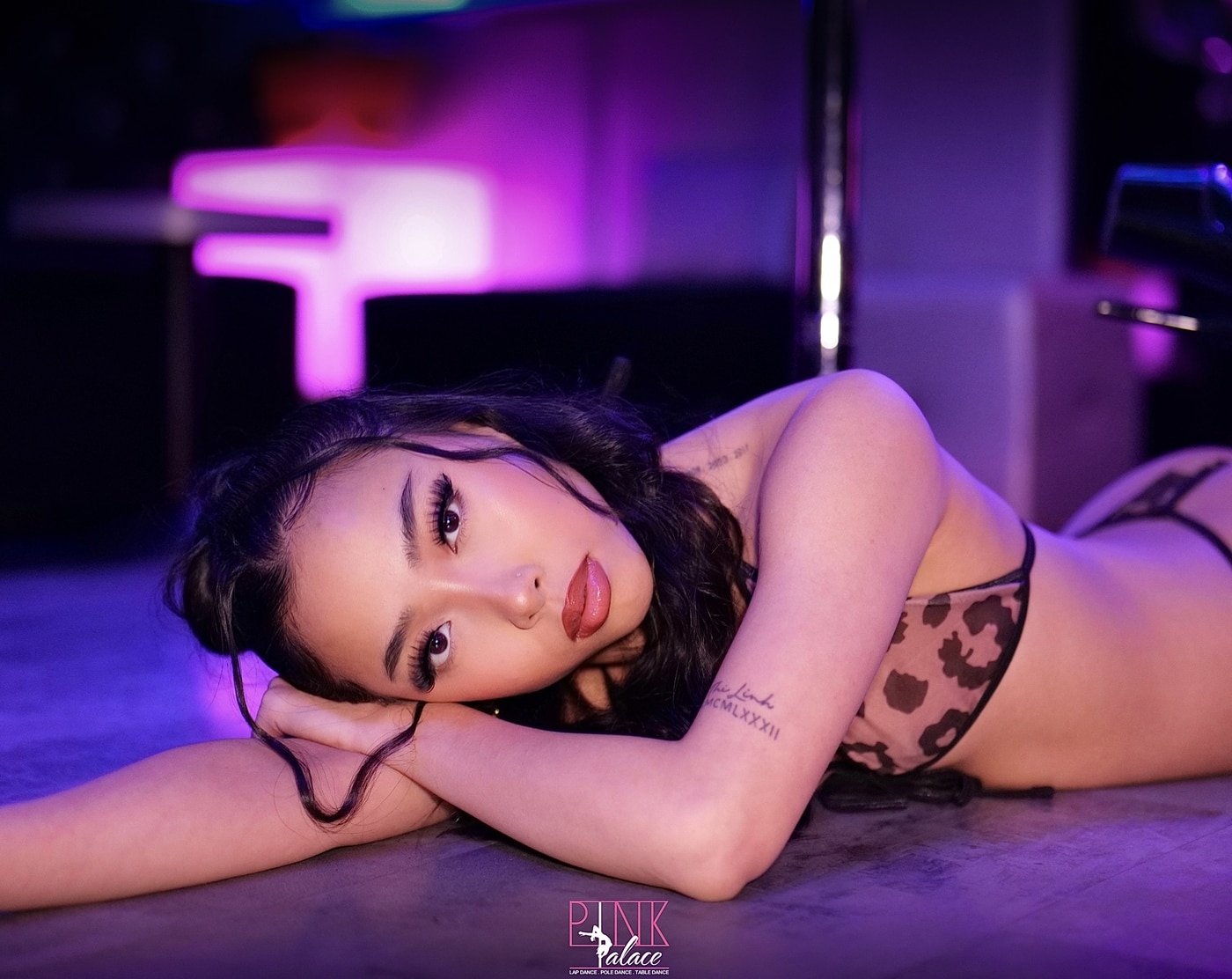 Jeune femme brune en soutien-gorge léopard se repose sur le sol, éclairage rose/violet. Tatouage sur le bras, logo "Pink Palace".