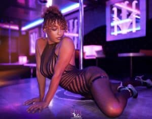 Danseuse en body résille et collants à fishnet, allongée au sol près d'une barre de pole dance, éclairage violet/rose. Logo "Pink Palace".