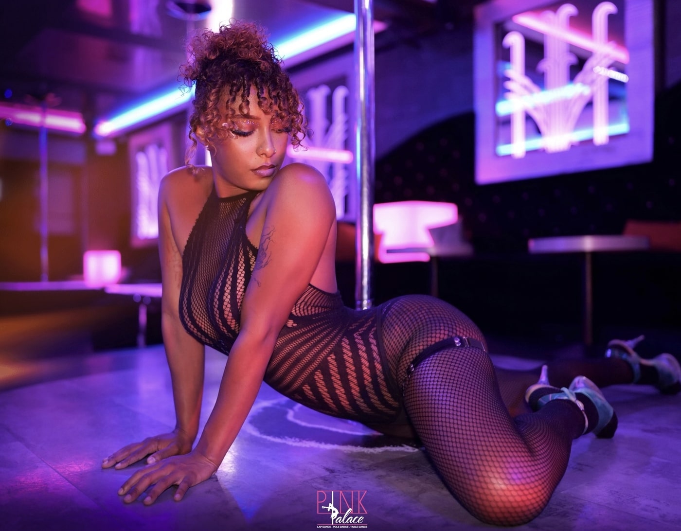 Danseuse en body résille et collants à fishnet, allongée au sol près d'une barre de pole dance, éclairage violet/rose. Logo "Pink Palace".