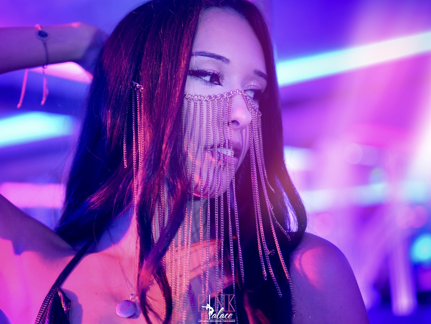 Jeune femme aux longs cheveux noirs portant un masque en chaînes dorées, éclairée par des lumières magenta et bleu vif, avec le logo Pink Palace.