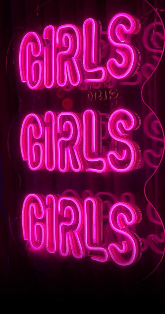 Enseigne néon rose vif répétant GIRLS trois fois verticalement sur un fond sombre, ambiance urbaine rétro.